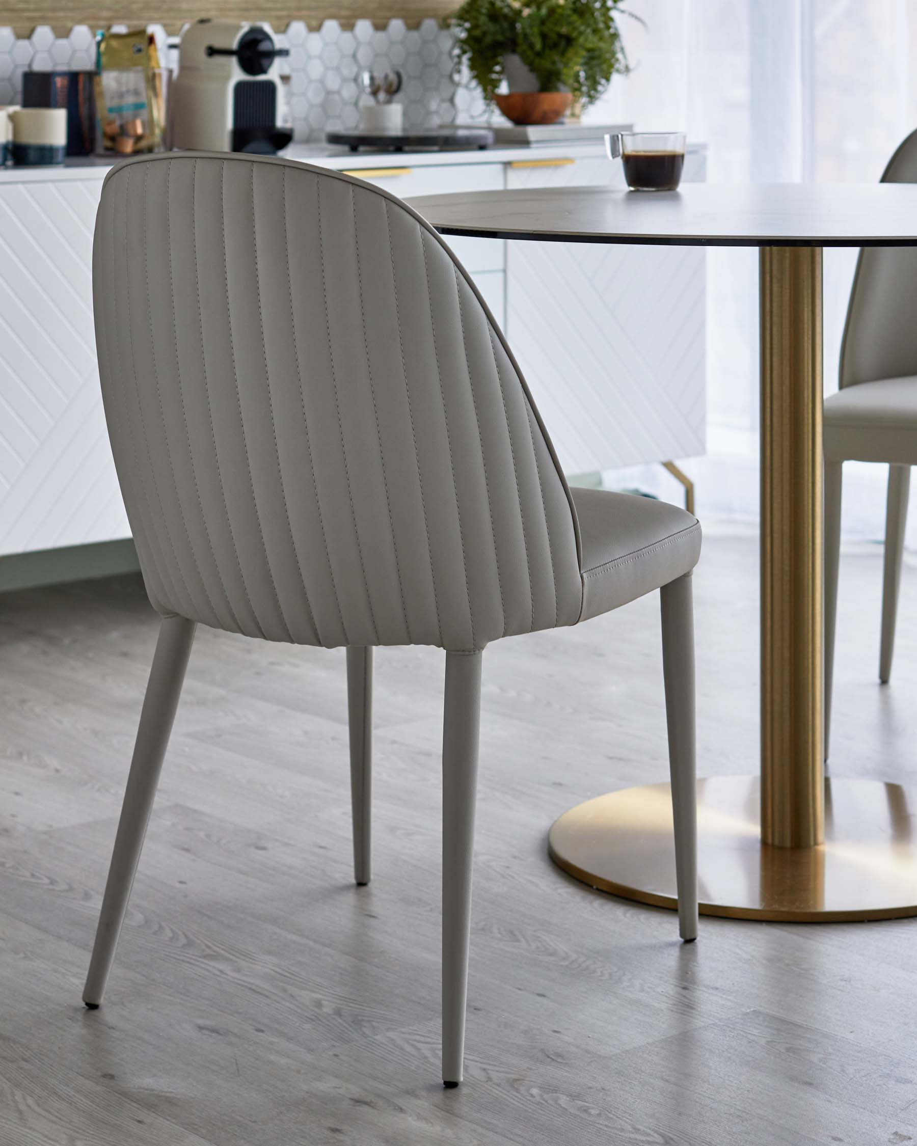 A stylish, upholstered beige dining chair with vertical stitching and slender legs, paired with a round table featuring a gold base.