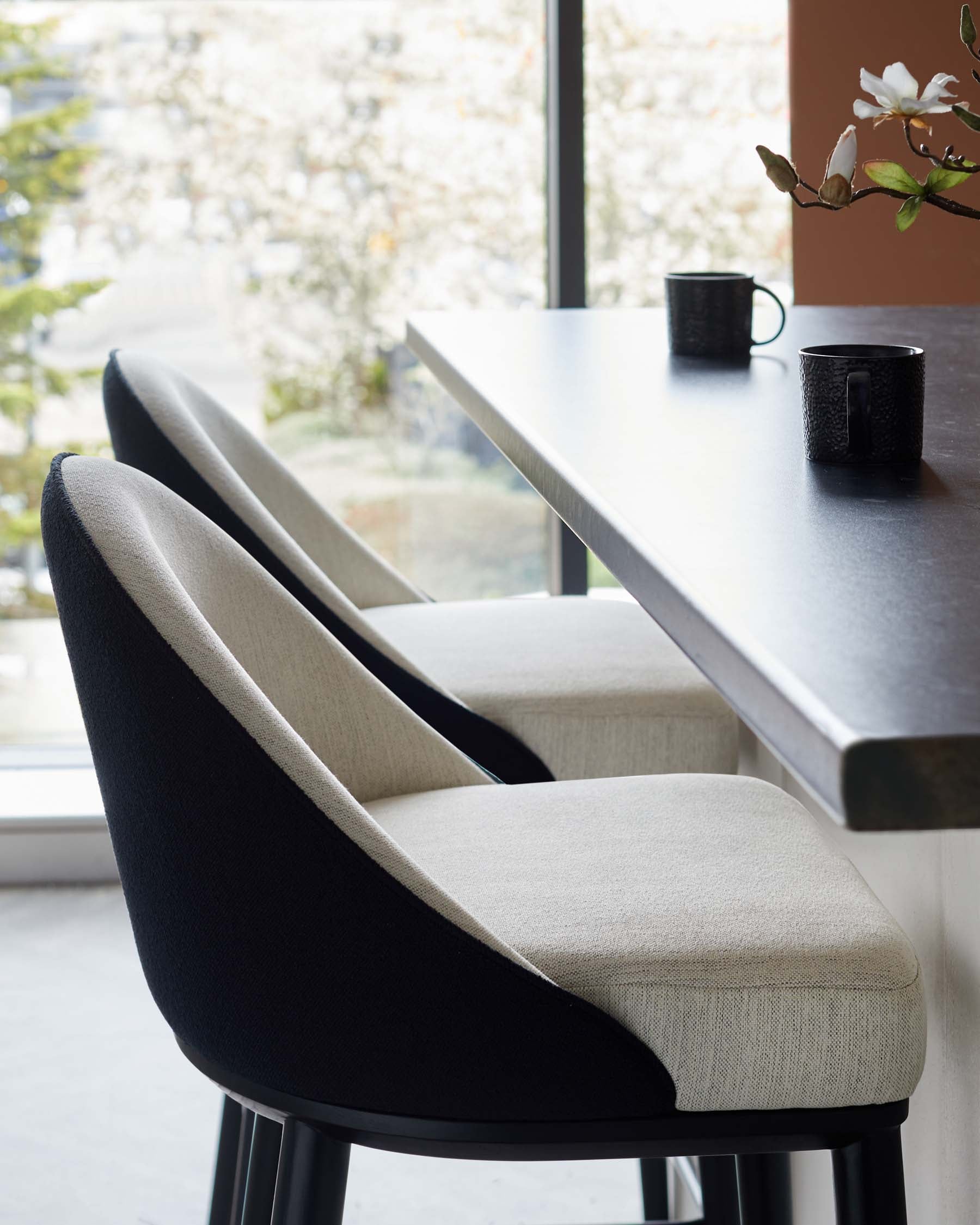 Two elegant bar stools with a mix of black and beige upholstery, positioned beside a sleek wooden table, complemented by dark mugs.