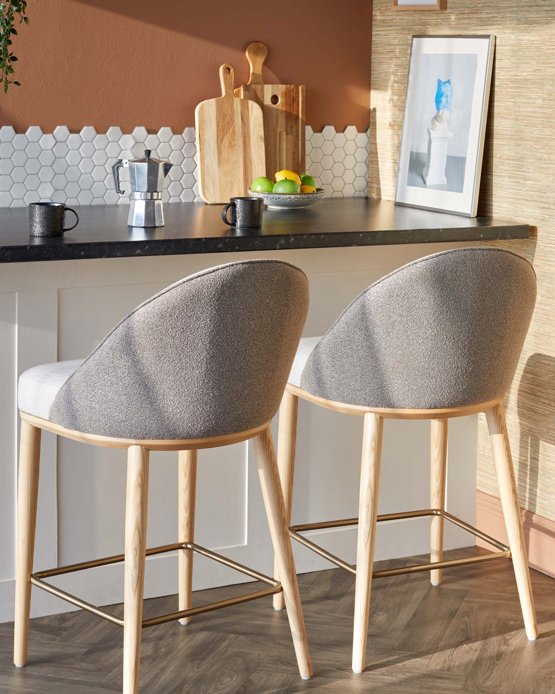 Two modern bar stools with gray upholstery and wooden legs, positioned at a sleek kitchen counter alongside coffee cups and decor.