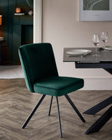 Elegant modern dining chair with plush green velvet upholstery and sleek black metal legs, accompanied by a contemporary black dining table with a clear set tableware arrangement, against a background featuring a subdued colour palette and minimalist decor.