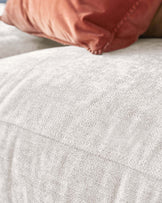 Close-up view of a textured light grey sofa with a plush surface, accented by a soft pink pillow with a textured fringe edge.