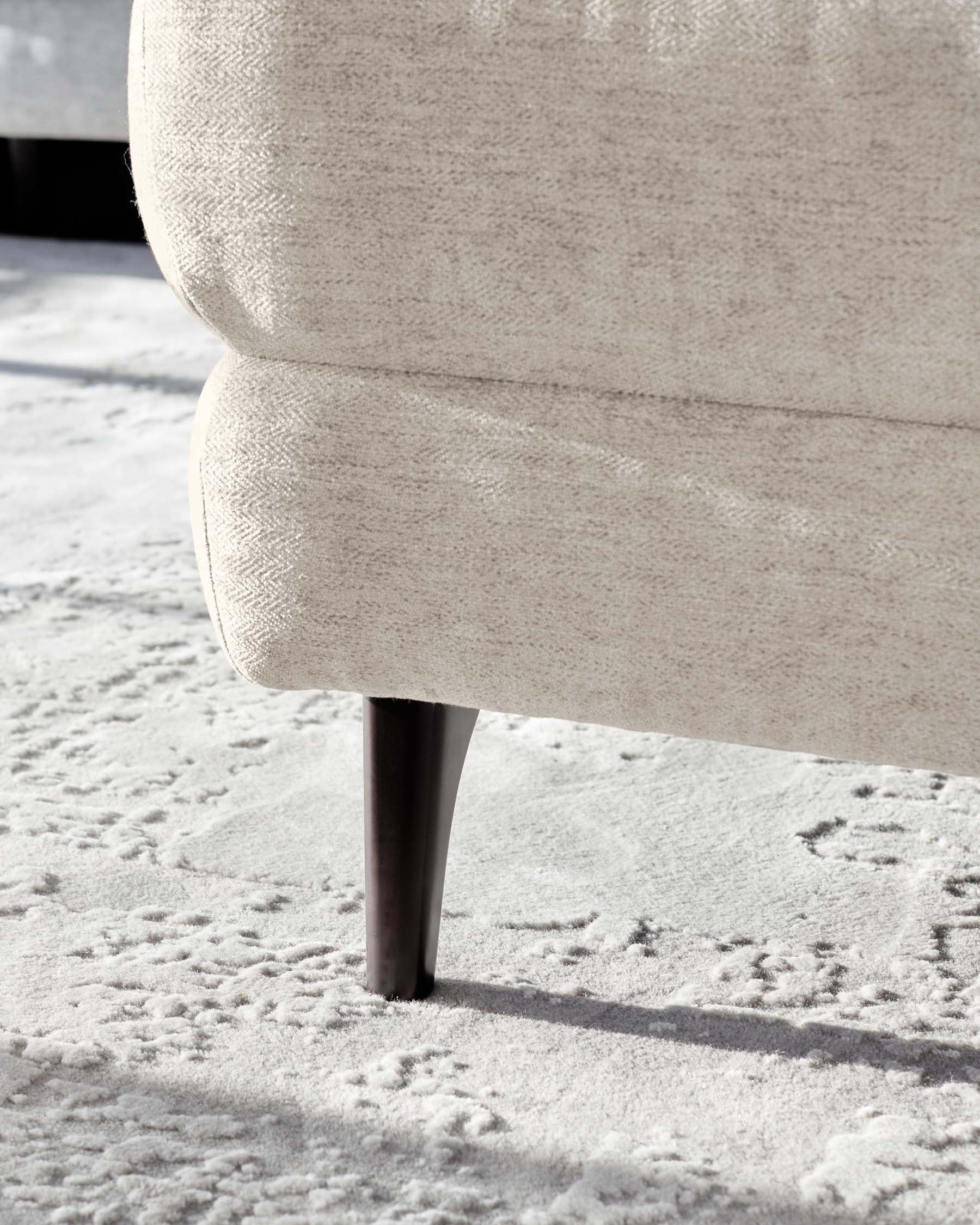 Close-up view of a cream-colored textured fabric sofa with a tapered dark wooden leg, placed on a white patterned area rug.