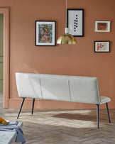 A sleek white bench with a minimalist design and black legs, set against a warm terracotta wall with stylish artwork.