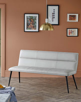 A sleek, white upholstered bench with a minimalist design, featuring black legs, set against a warm brown wall with artwork.