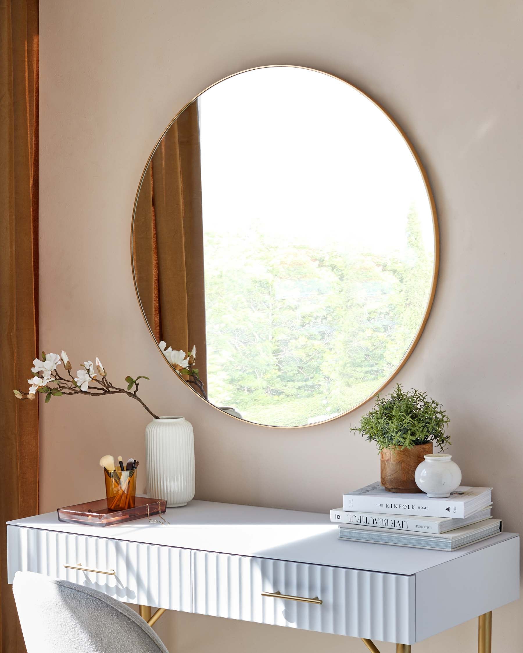 A contemporary white desk with a textured front design and elegant brass handles, paired with a large round mirror featuring a thin gold frame. The wall behind is a soft peach colour, and the desk is accessorized with a white vase, a small green plant, a stack of books, and a clear makeup organizer with brushes.