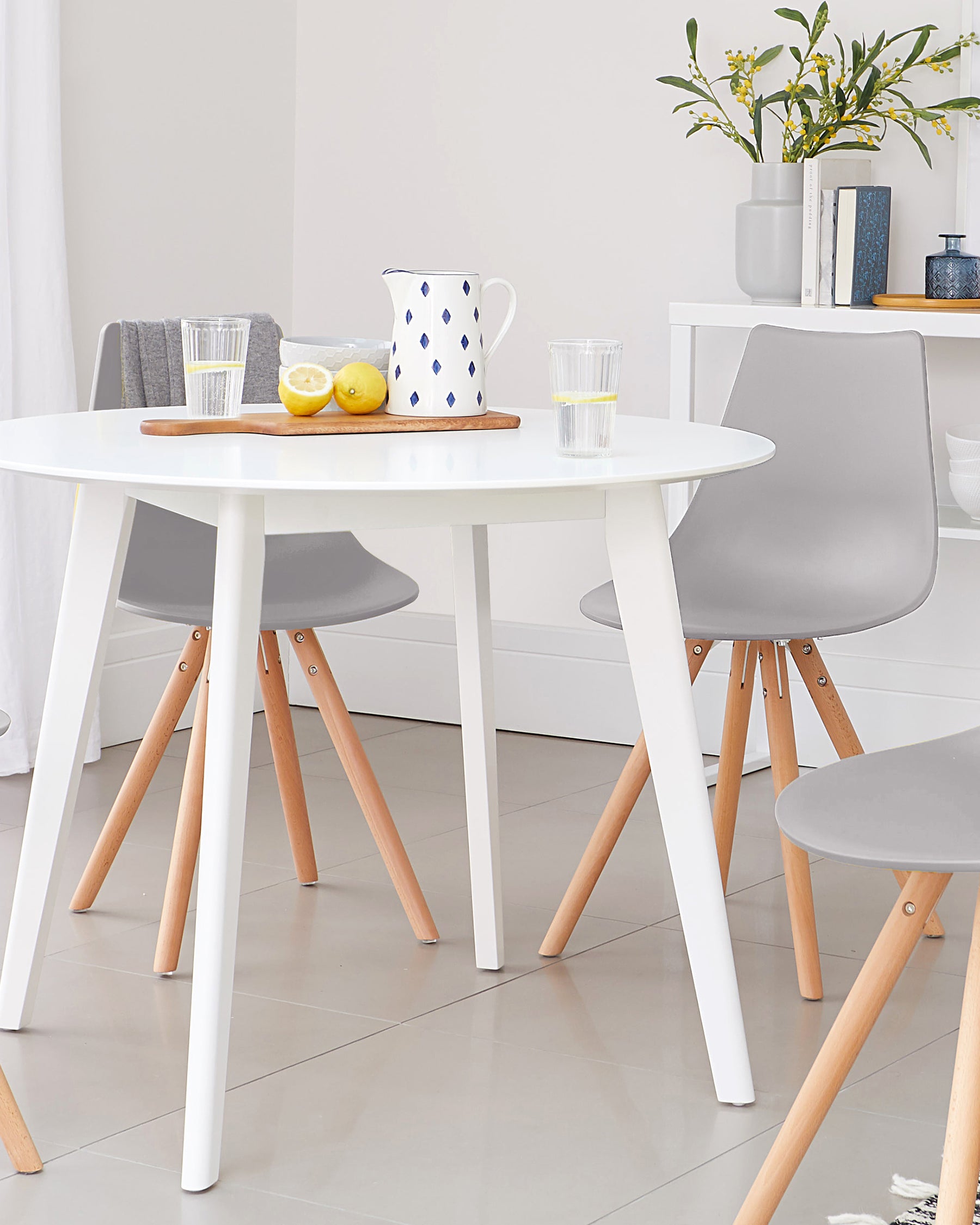 A round white table with wooden legs, paired with gray chairs featuring wooden accents, set with a pitcher and glasses.