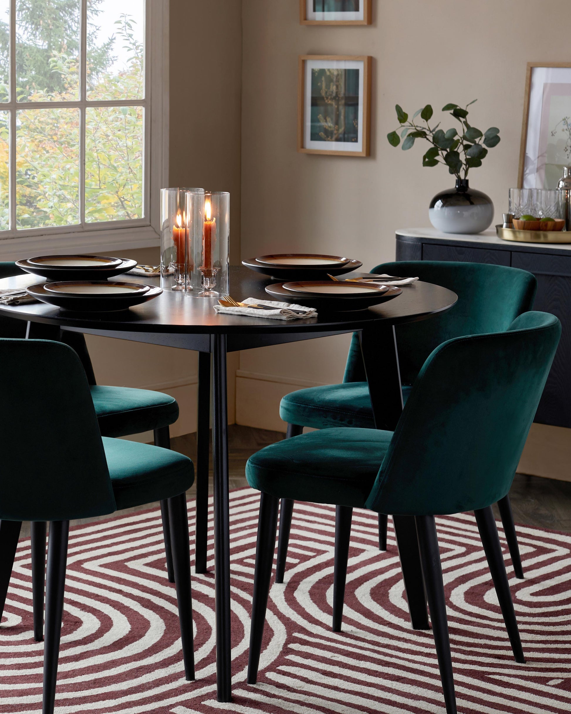 A round black dining table with four green velvet chairs, set elegantly with plates and candle holders on a patterned rug.