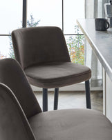 Modern minimalist dining chair with a sleek dark grey velvet upholstery and sturdy metal legs in a deep blue tone, placed next to a white marble countertop.