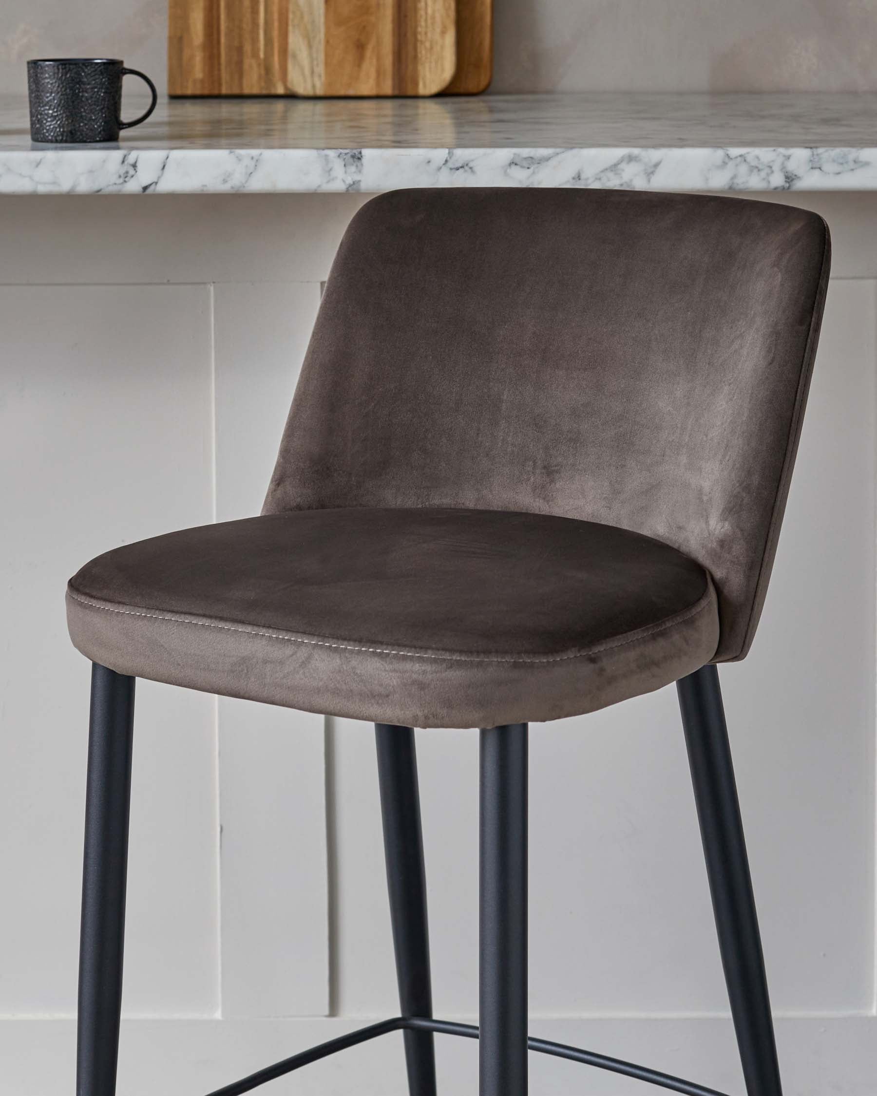 Modern charcoal grey velvet bar stool with a curved backrest and cushioned seat, mounted on a sleek black metal frame with four slender legs and geometric footrests. The stool is positioned against a kitchen island with a white marble countertop.