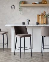 Elegant modern kitchen with a minimalist design featuring a sleek white marble countertop and two luxurious velvety brown bar stools with slender black metal legs set against a textured grey wall. The barstools have a contemporary design with a subtly curved backrest and a comfortable footrest.