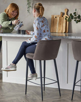 Two modern bar stools with sleek taupe upholstery and thin black metal legs, featuring a curved backrest and comfortable seat design for contemporary kitchen or bar settings.