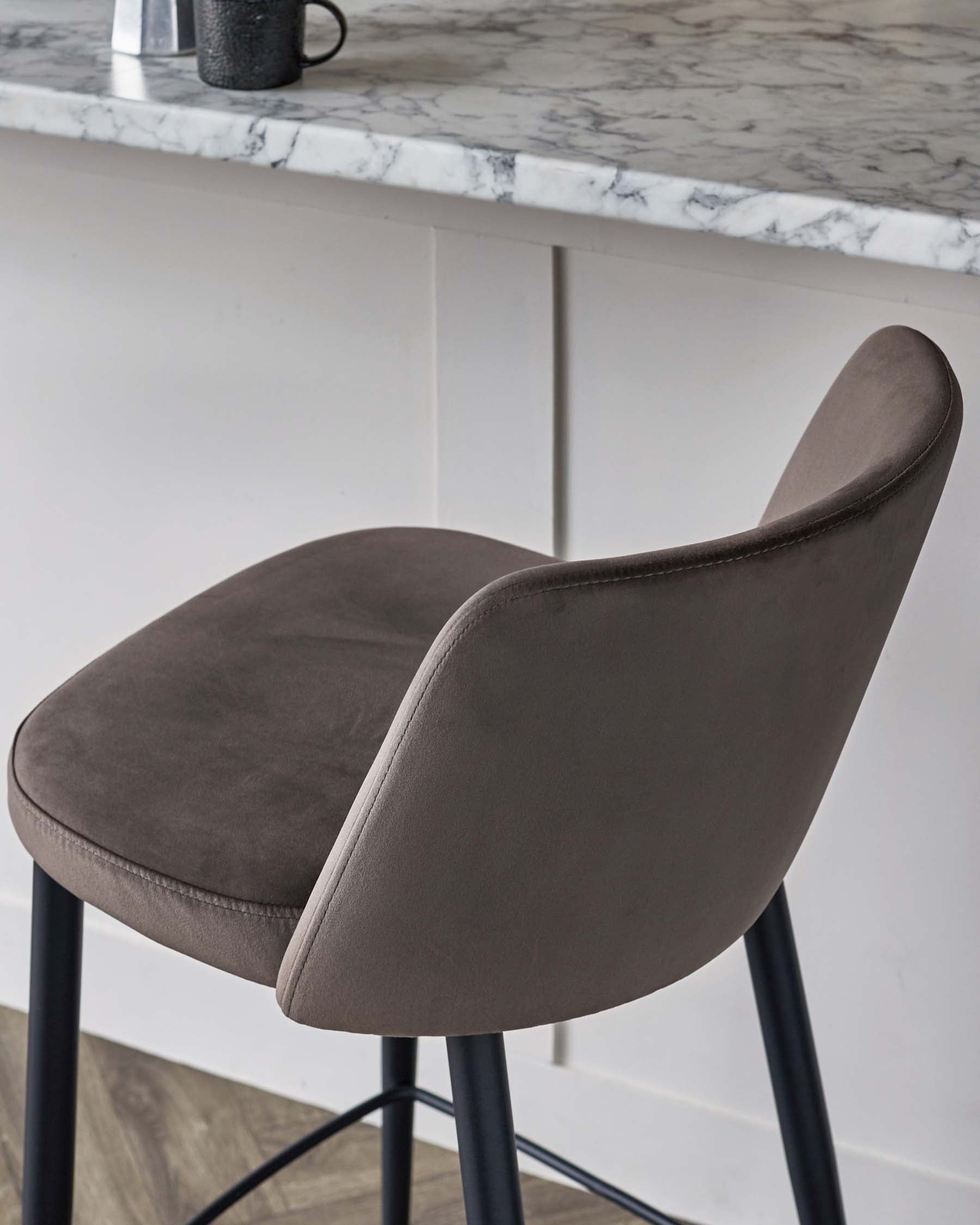 Modern grey upholstered bar stool with a curved backrest and sleek black metal legs, positioned up against a white and grey marble countertop.