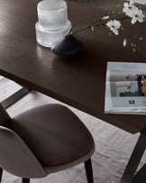 Modern minimalist furniture setup featuring a sleek, dark wooden table with angular metal legs, complemented by a plush, brown fabric-upholstered dining chair with a curved backrest and black metal legs. A decorative vase and a sprig with white blossoms subtly adorn the table, suggesting an elegant and contemporary dining ambiance.