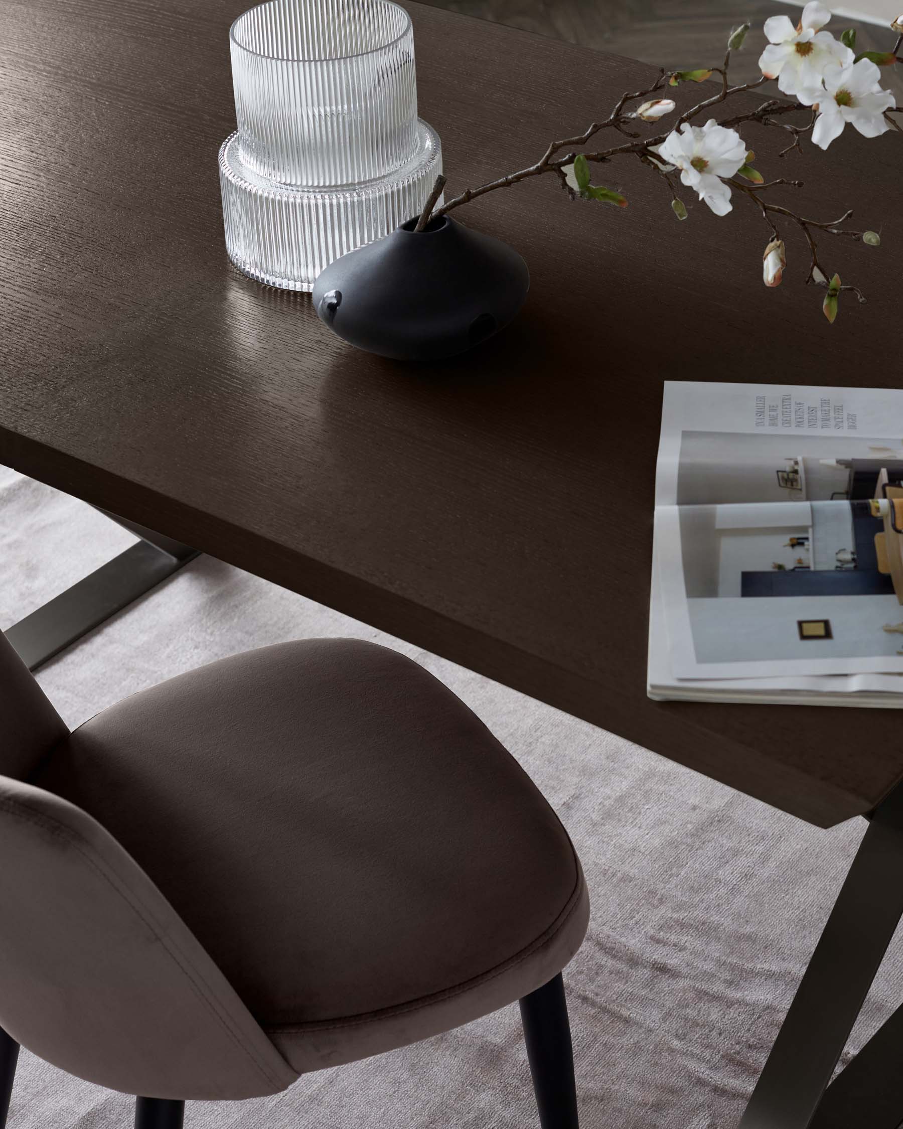 Modern minimalist furniture setup featuring a sleek, dark wooden table with angular metal legs, complemented by a plush, brown fabric-upholstered dining chair with a curved backrest and black metal legs. A decorative vase and a sprig with white blossoms subtly adorn the table, suggesting an elegant and contemporary dining ambiance.