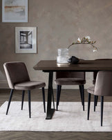 A contemporary dining set featuring a dark wooden table with unique angled black metal legs and three matching velvet-upholstered chairs with slender black legs. The set is arranged on a pale textured area rug.