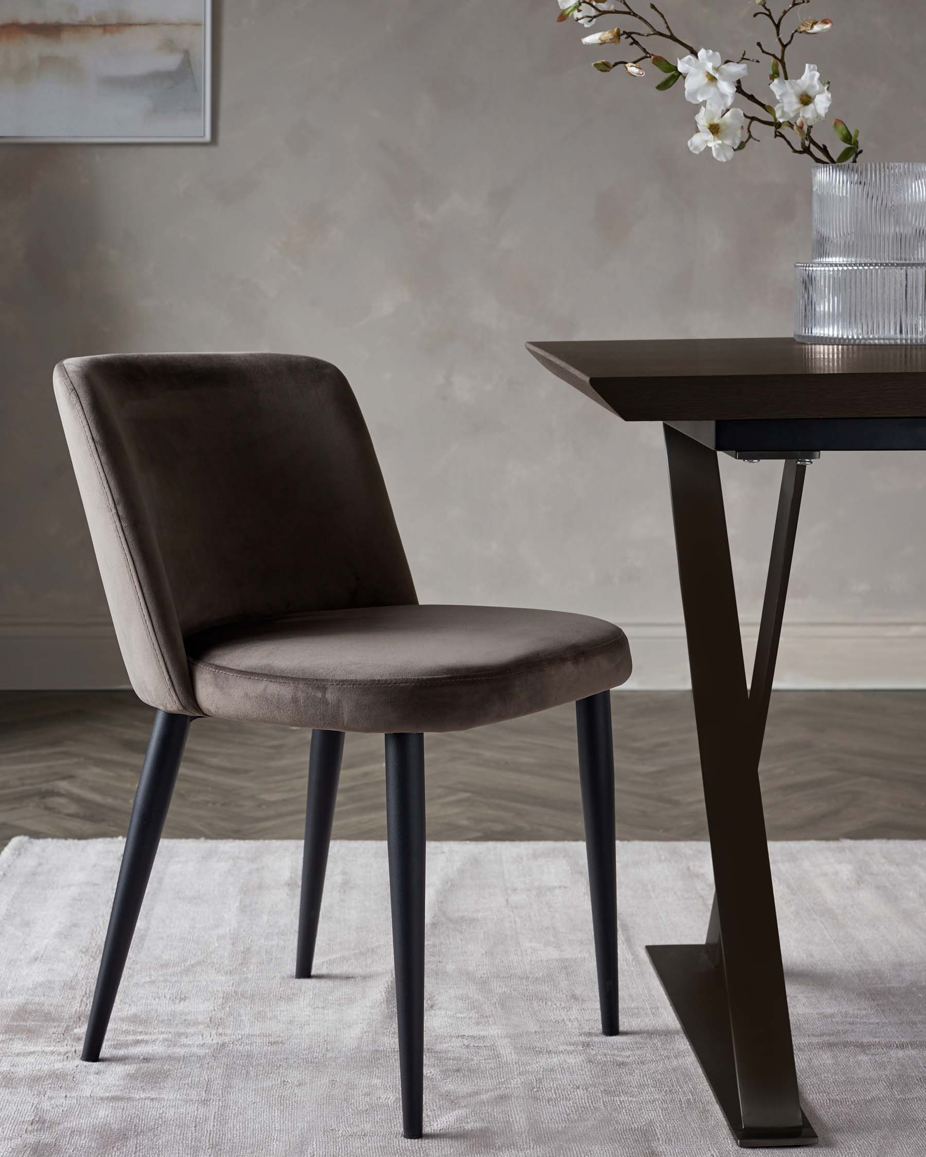 Sleek gray velvet chair with black legs beside a modern table featuring a geometric base, complemented by a decorative vase.