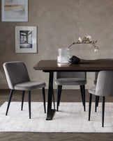 A modern minimalist dining set featuring a rectangular table with a dark wood finish and angular black metal legs, accompanied by three grey upholstered chairs with black tapered legs, set on a white textured area rug.