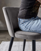 Modern grey velvet dining chair with a curved backrest and cushioned seating, standing on sleek black metal legs.