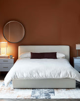 A modern bed with a light fabric headboard, white bedding, paired with two nightstands and stylish lamps, on a colorful rug.