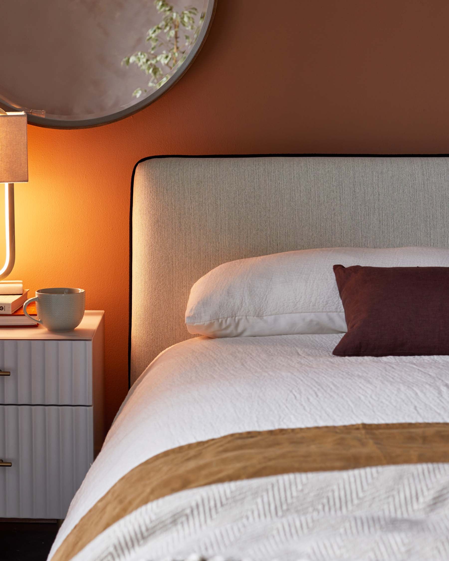 A cozy bedroom featuring a tufted headboard, a textured white duvet, a stylish nightstand with books, a lamp, and a mug.