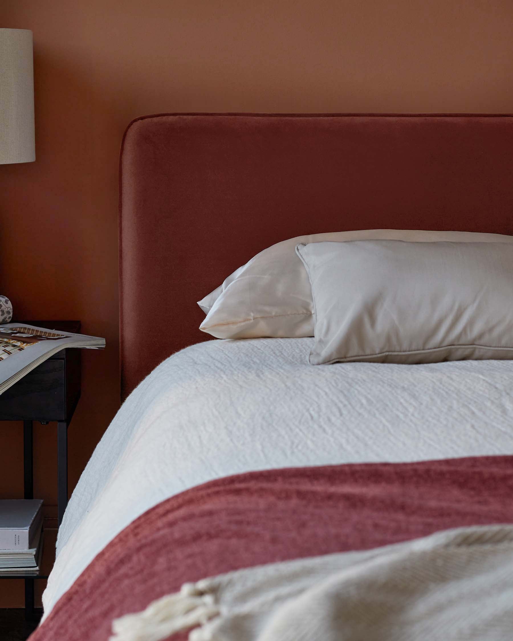 Cozy bedroom featuring a plush rust-colored headboard, soft white bedding, and a bedside table for magazines and decor.