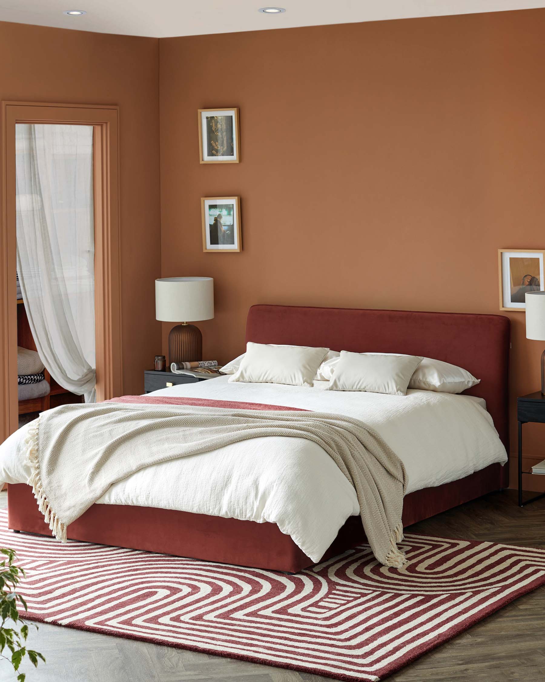 A modern bedroom featuring a burgundy bed, white bedding, textured blanket, and a patterned area rug against a warm-toned wall.