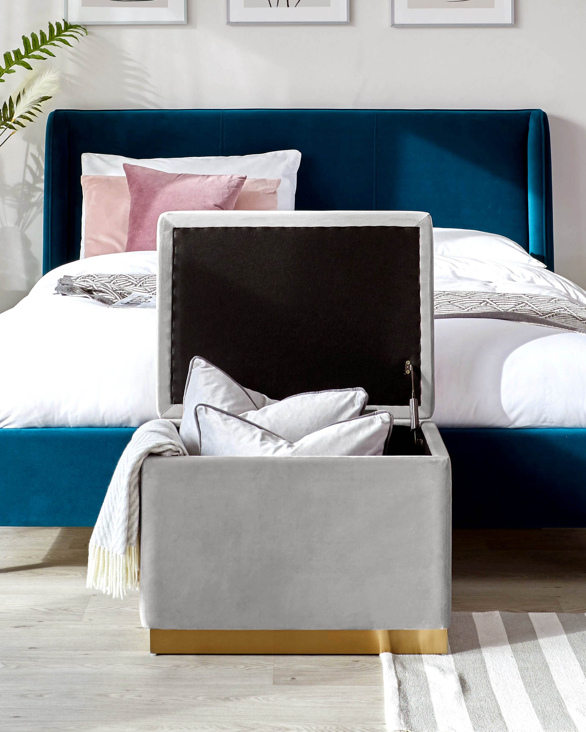 Stylish gray storage ottoman with gold accents, complemented by a plush blue bed and decorative pillows in a modern bedroom setting.