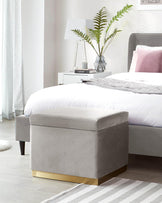 Gray velvet storage ottoman with a gold base, placed beside a bed in a modern bedroom setting.