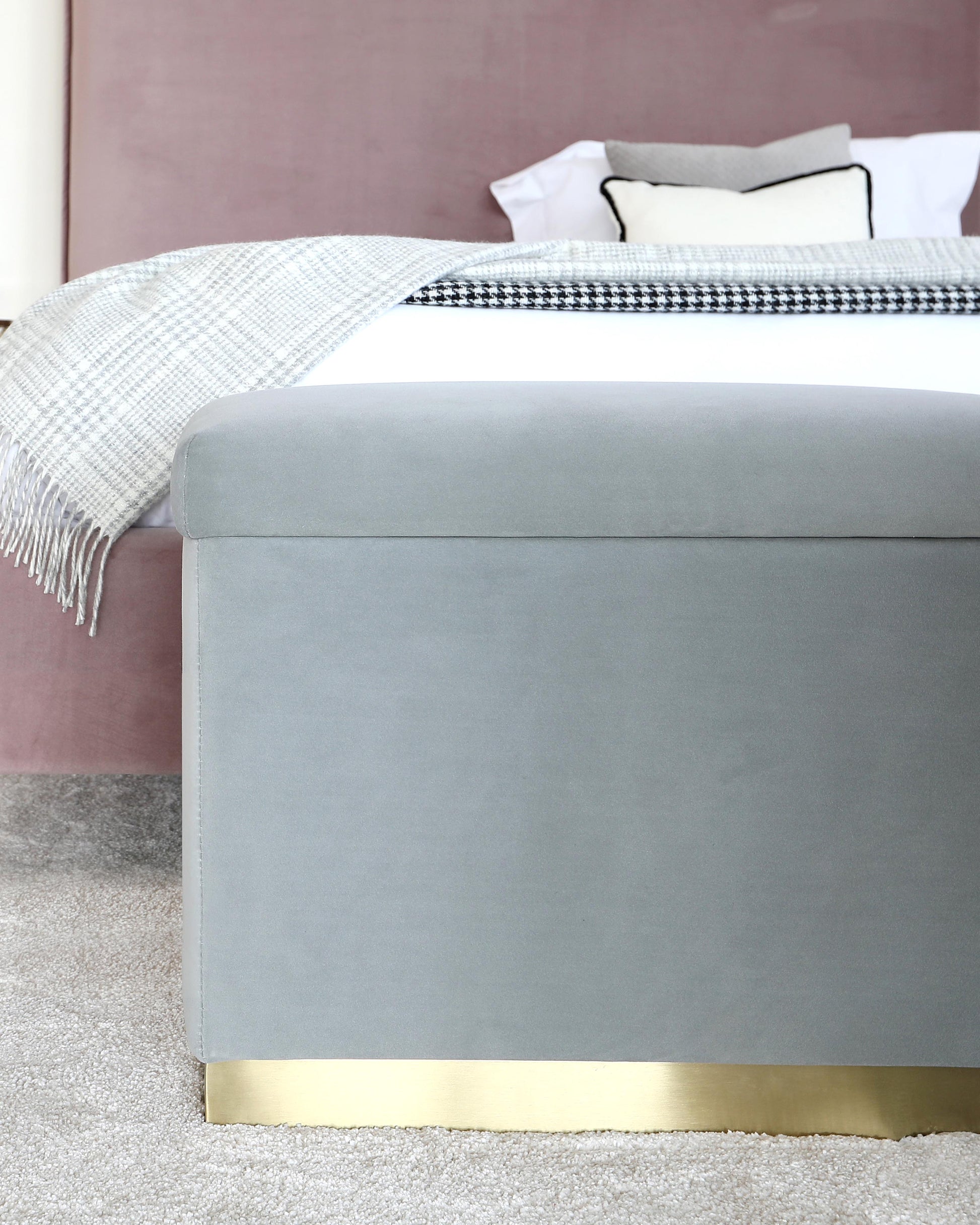 A soft gray ottoman with a sleek gold base, placed beside a bed with a textured throw blanket and decorative pillows.