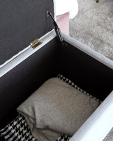 A light gray storage ottoman with a soft interior, holding a folded sweater and a patterned throw blanket inside.