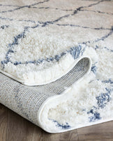 Plush white and blue patterned area rug with a high-pile texture, partially rolled up to reveal its underside on a wooden floor.