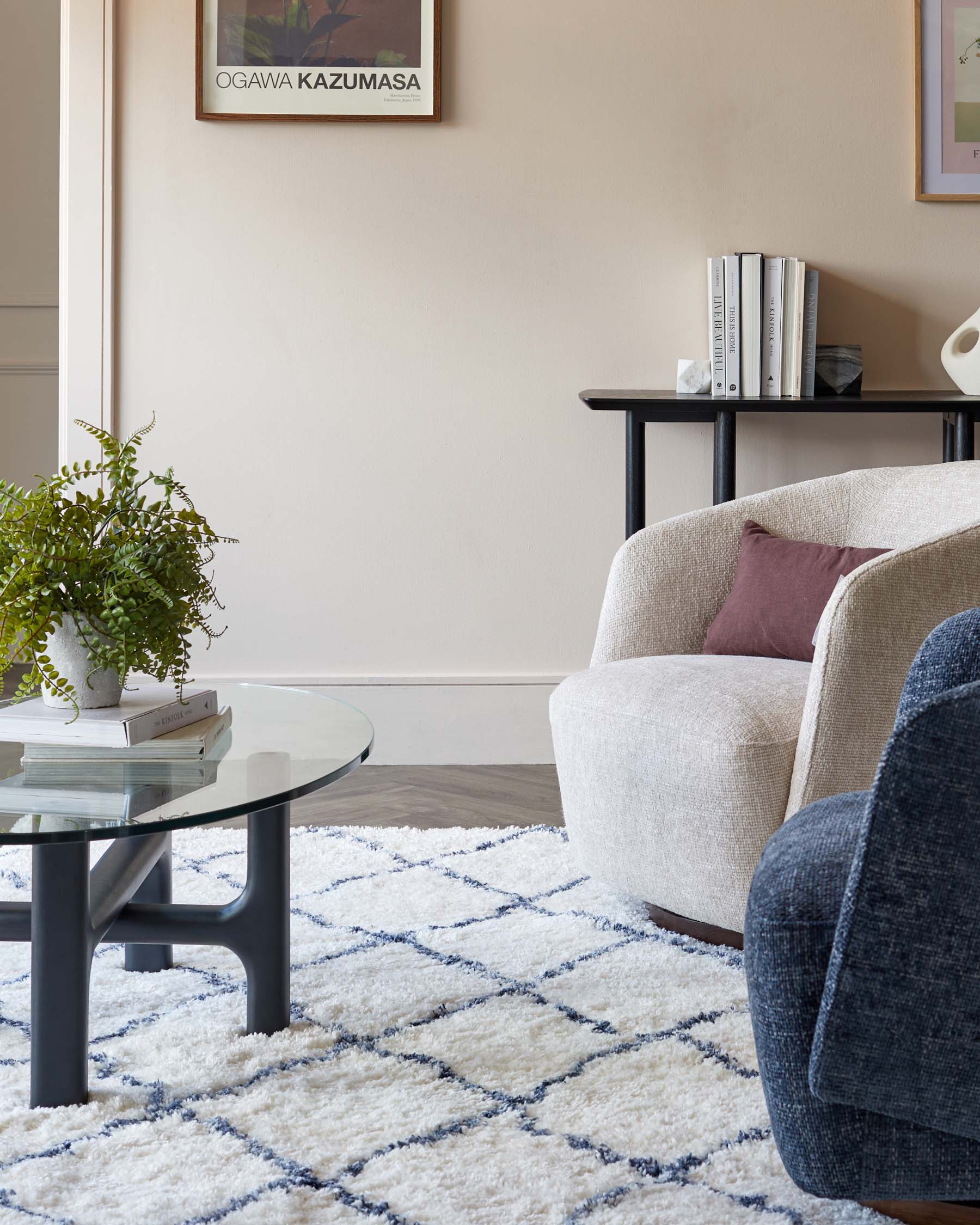 A cosy modern living room setup featuring a cream textured fabric armchair with a dark purple throw pillow, a round glass coffee table with a black matte metal frame, and a sleek black console table holding books and decorative items. The ensemble is complemented by a plush white rug with blue geometric patterns.