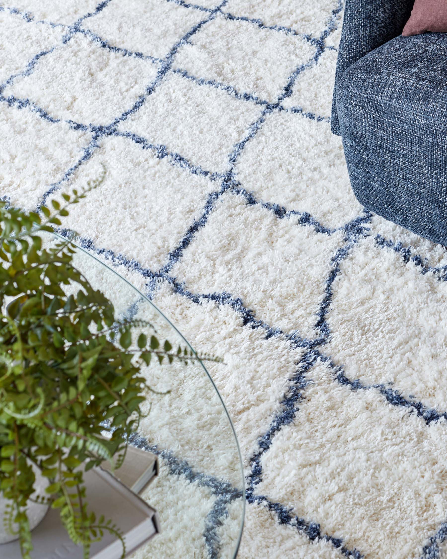 Plush off-white area rug with a diamond pattern in blue, partially under a grey upholstered chair with a pink pillow. A round glass side table with a plant and books is also visible.