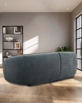 A curved, plush teal sofa and a modern black bookshelf showcase stylish furniture in a sunlit room with wooden flooring.