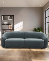 Curved teal sofa with soft fabric, two-seater design, complemented by a modern open shelf unit in a bright, airy room.