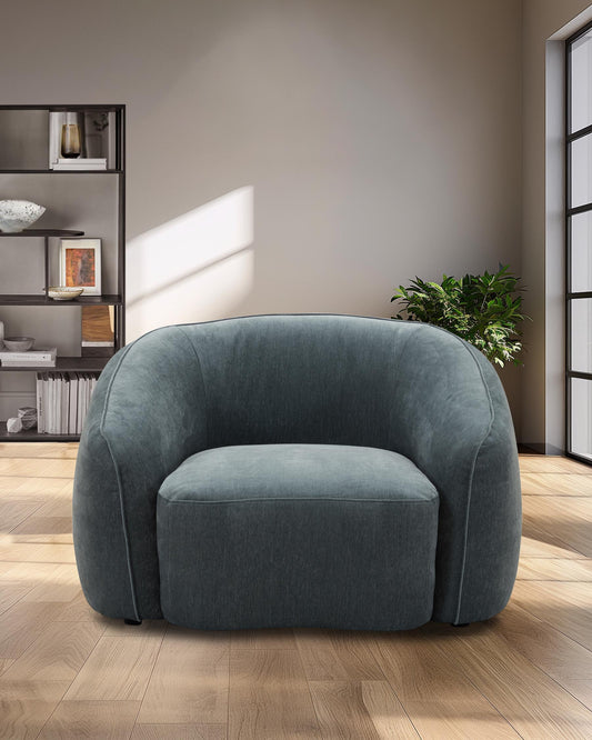 A stylish, rounded armchair in a rich teal fabric, featuring a cozy cushion and paired with modern shelving in a bright space.