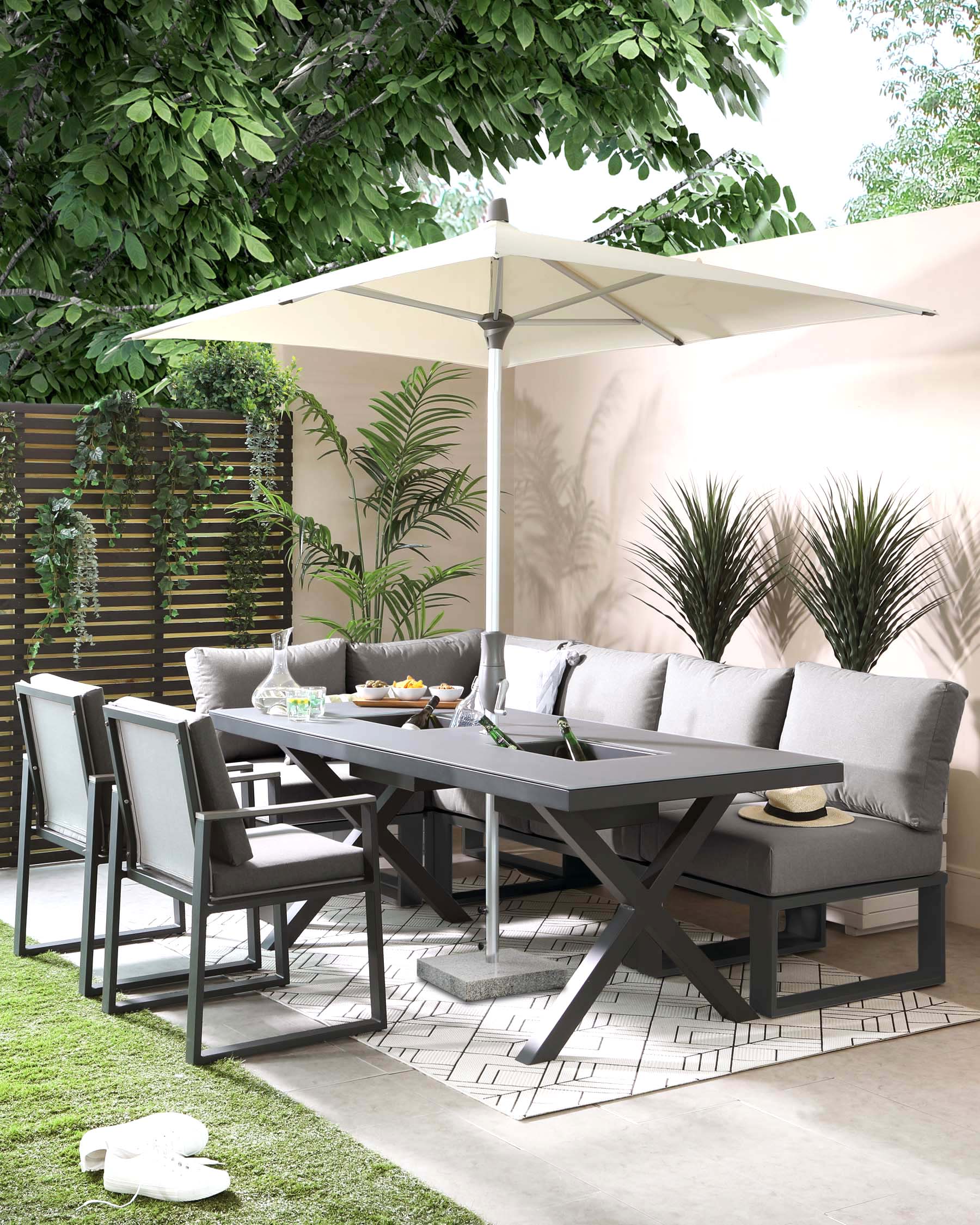 Outdoor dining furniture set under a patio umbrella featuring a grey rectangular table with an X-base design, surrounded by six armchairs with fabric cushions, and a matching grey outdoor sofa with white accent pillows, all set against a backdrop of lush greenery and privacy fencing.