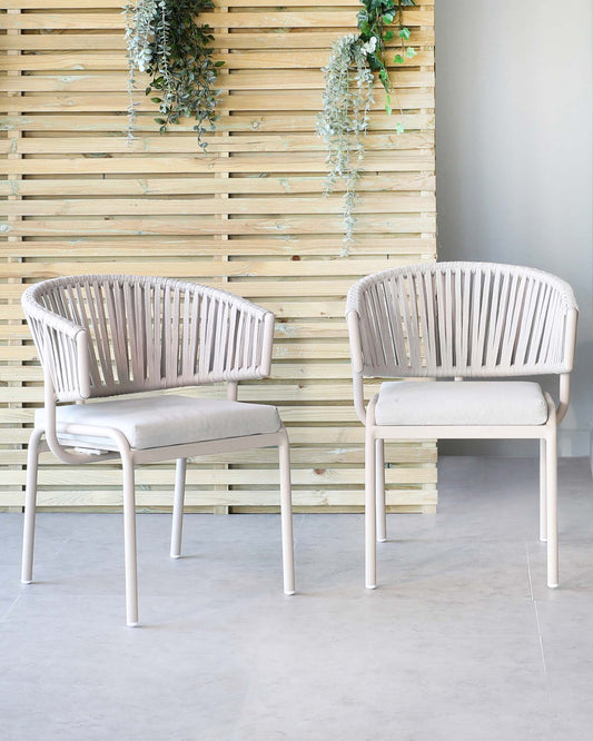 Two contemporary light beige armchairs with a vertical slat back design and upholstered seat pads, set against a wooden slat wall with hanging greenery.