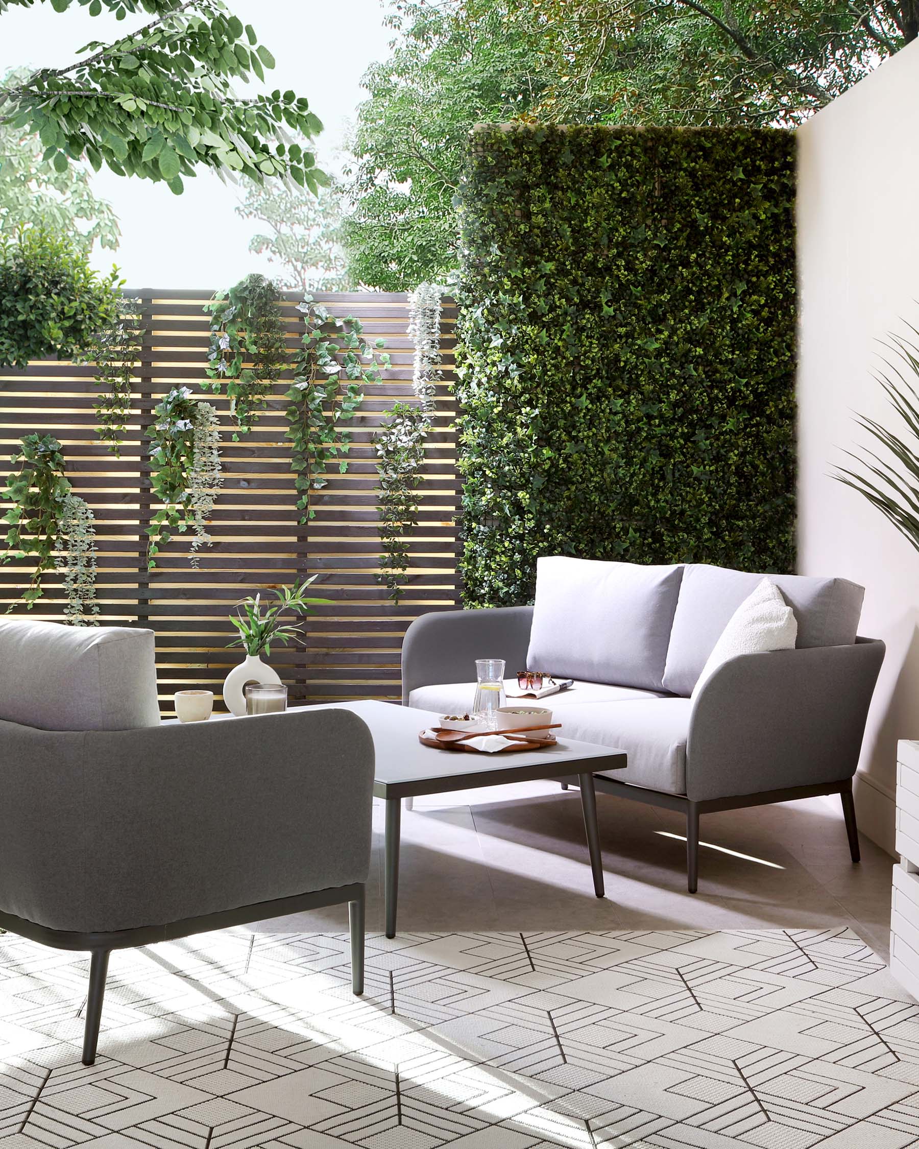 A modern outdoor patio furniture set that includes a dark grey three-seater sofa with pillows and two matching armchairs, all with sleek wooden legs, accompanied by a rectangular coffee table in a matching finish. The set is arranged on a tiled patio with geometric patterns, enclosed by greenery and wooden slat fencing.