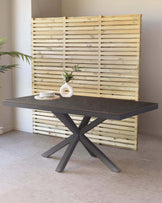 A modern dining table with a dark grey stone-like tabletop resting on a striking matte black double X-shaped metal base. The table is set against a natural wood slat backdrop, accompanied by a small round white vase with green foliage on its surface, enhancing its contemporary design.