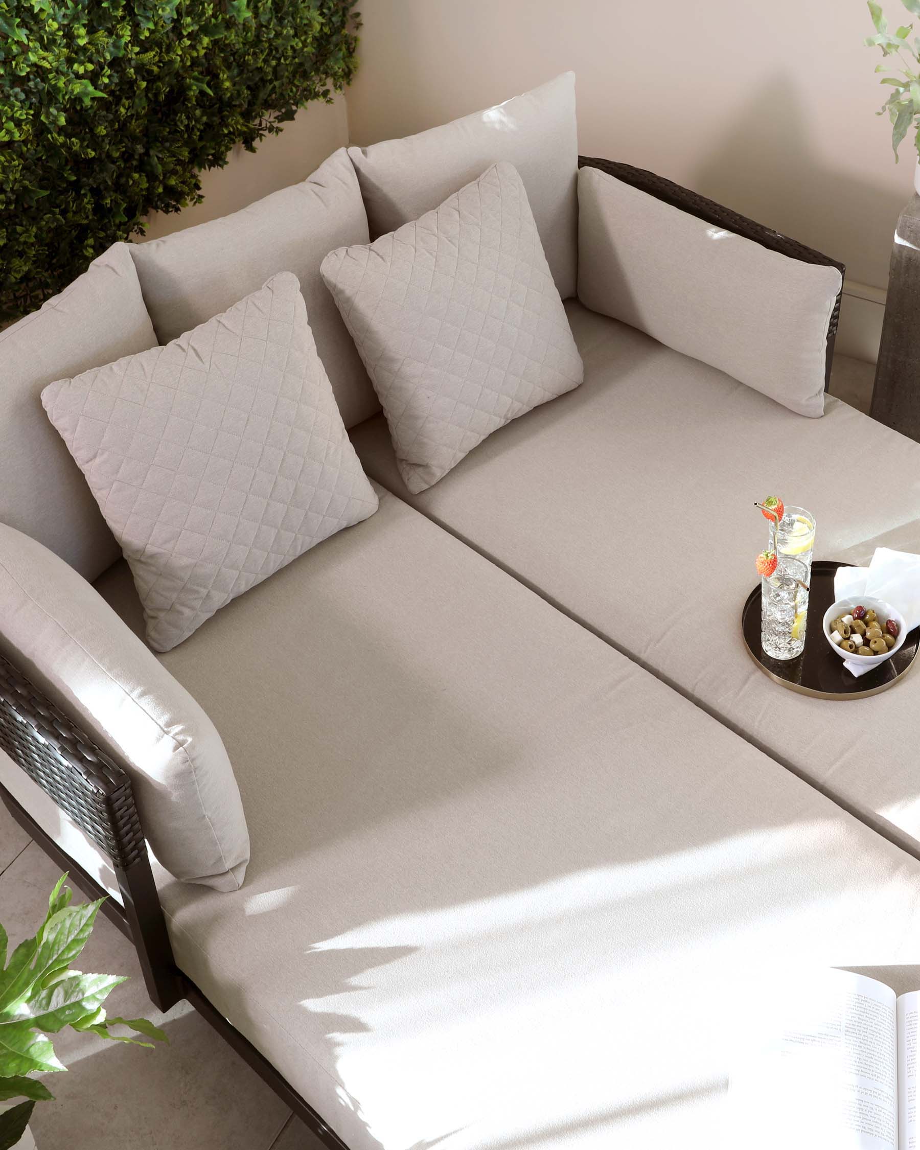 Contemporary outdoor L-shaped sectional sofa with a beige upholstered finish and matching quilted square throw pillows, featuring a dark wicker accent on the sofa's side and armrests. A small round side table with a black tray holding a glass of beverage and a bowl of olives is placed on the sofa. The setting is complemented by green foliage and sunlight casting soft shadows, evoking a calm and inviting outdoor atmosphere.