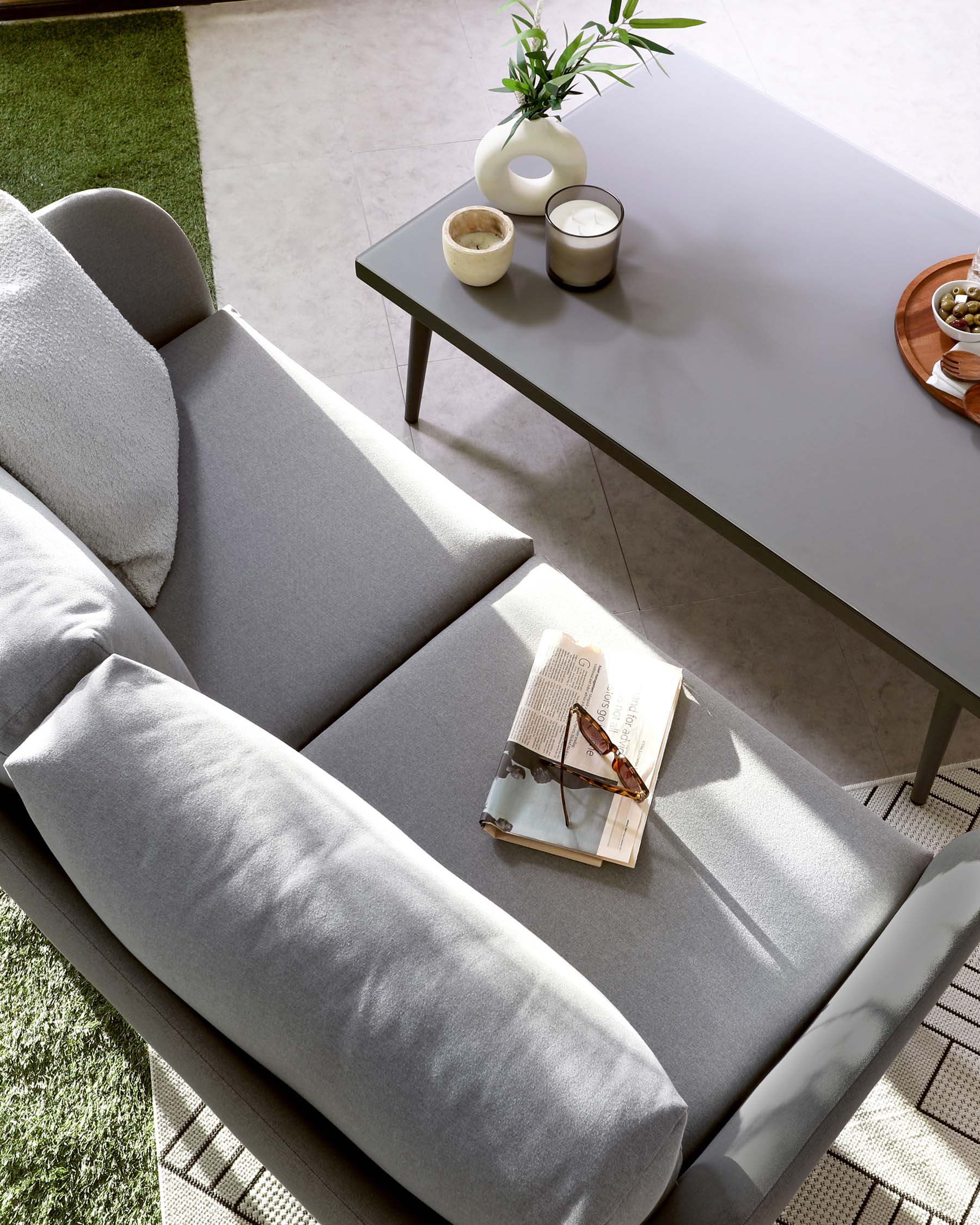 Modern minimalist furniture scene featuring a light grey upholstered sofa with rounded edges and a cylindrical bolster cushion, positioned next to a low-profile grey rectangular coffee table adorned with simple white ceramic vases, candles, and a wooden tray with snacks. The setting is complemented by a textured rug and a hint of greenery, with natural light casting soft shadows across the scene.