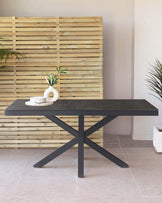 Modern minimalist dining table with a dark stone-like top and a stylish black X-shaped metal base, set against a backdrop of a natural wooden slat wall. The table is accessorized with a circular white vase containing green foliage and a stack of white ceramic plates on a woven placemat.