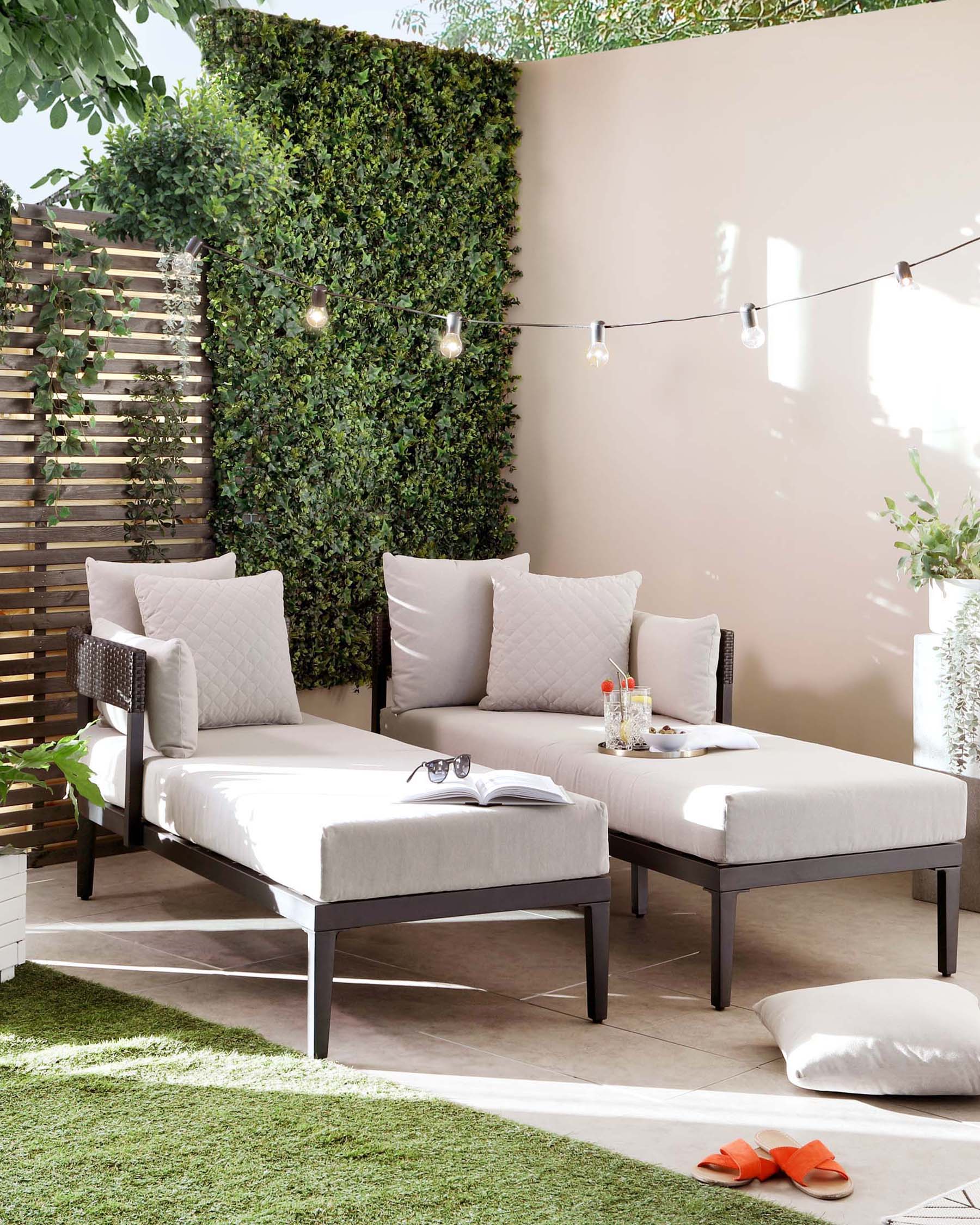 Contemporary outdoor furniture set featuring a pair of elegant chaise lounges with plush, light grey cushions and a sleek dark frame, complemented by a matching square side table with a clear glass top, arranged on a patio with decorative greenery and ambient string lights.