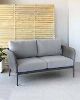 A modern two-seater sofa with grey upholstery and a sleek black metal frame, set against a wooden slat backdrop with a light finish. The sofa features clean lines and a minimalist design.