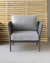 Modern single-seat armchair with grey upholstery, featuring a cushioned seat and backrest. The chair has a sleek, minimalist design with a matte black metal frame and legs, positioned against a wooden slat wall on a grey tile floor.