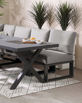 Modern grey upholstered corner bench with a matching contemporary dark grey dining table featuring a distinctive X-shaped base. The setting is complemented by a geometric patterned area rug below.