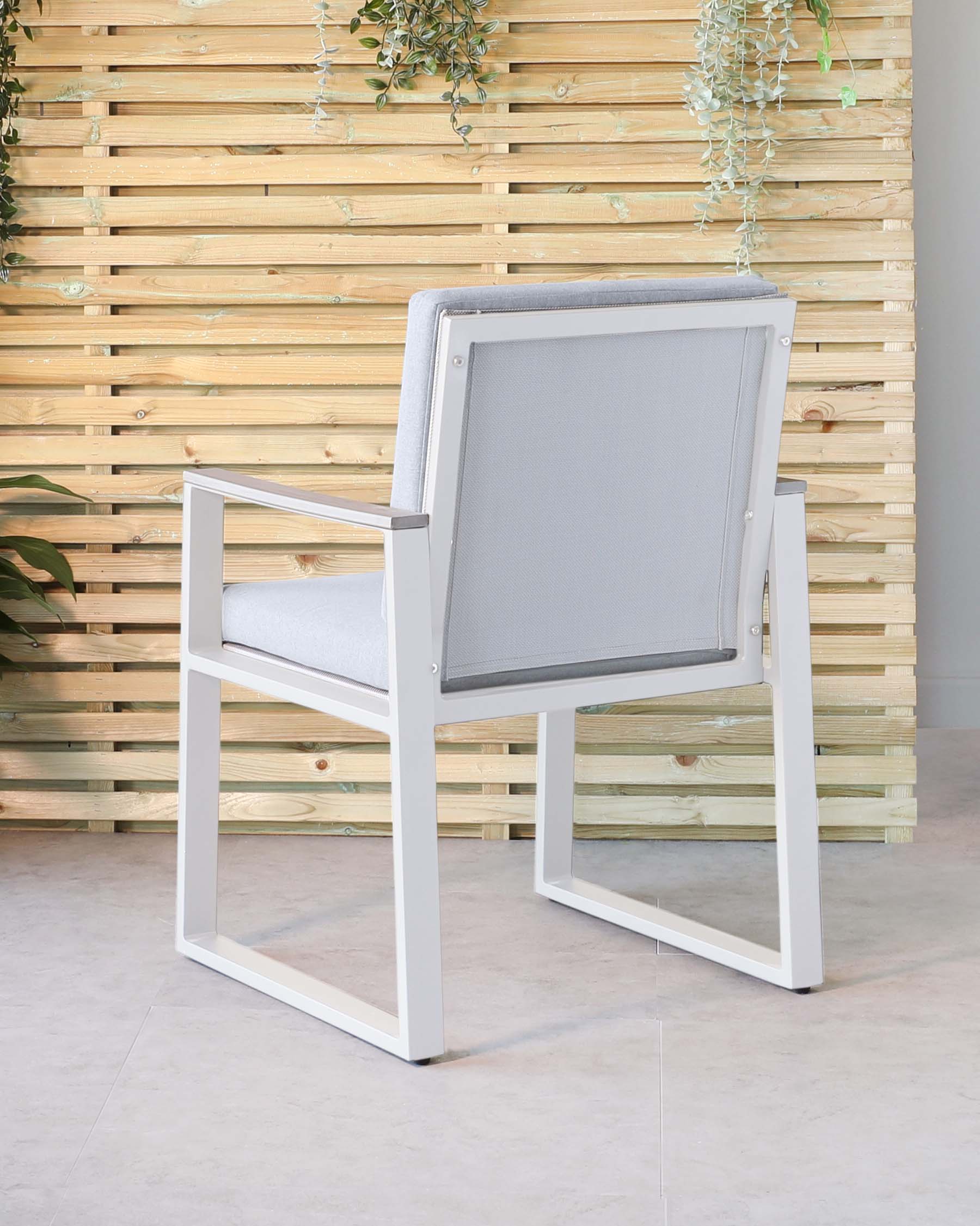 Modern outdoor armchair with a white aluminium frame, light grey waterproof fabric cushions, and a mesh backrest, standing against a wooden slat wall with green plants.