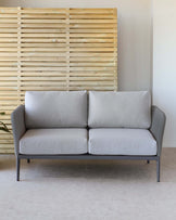 Modern two-seater sofa with light grey upholstery and a minimalist black metal frame, set against a wooden slat wall on a neutral-toned floor.