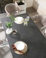 Modern outdoor dining set featuring a rectangular table with a dark grey matte top and a pair of elegant, curved, woven-back chairs in a natural beige tone. The chairs showcase an intricate vertical stripe pattern, providing both comfort and style for alfresco dining or patio lounging.